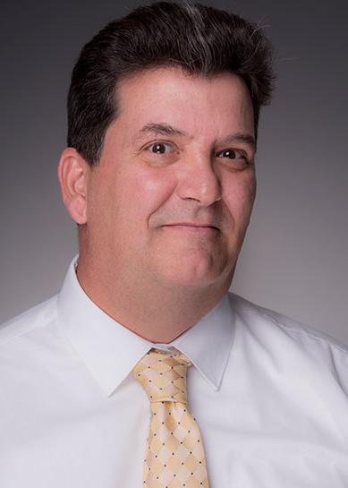 A Caucasian male with brown hair, Patrick Riley wears a white collared dress shirt and a gold diamond pattern tie with the diamond points separated by single black dots.